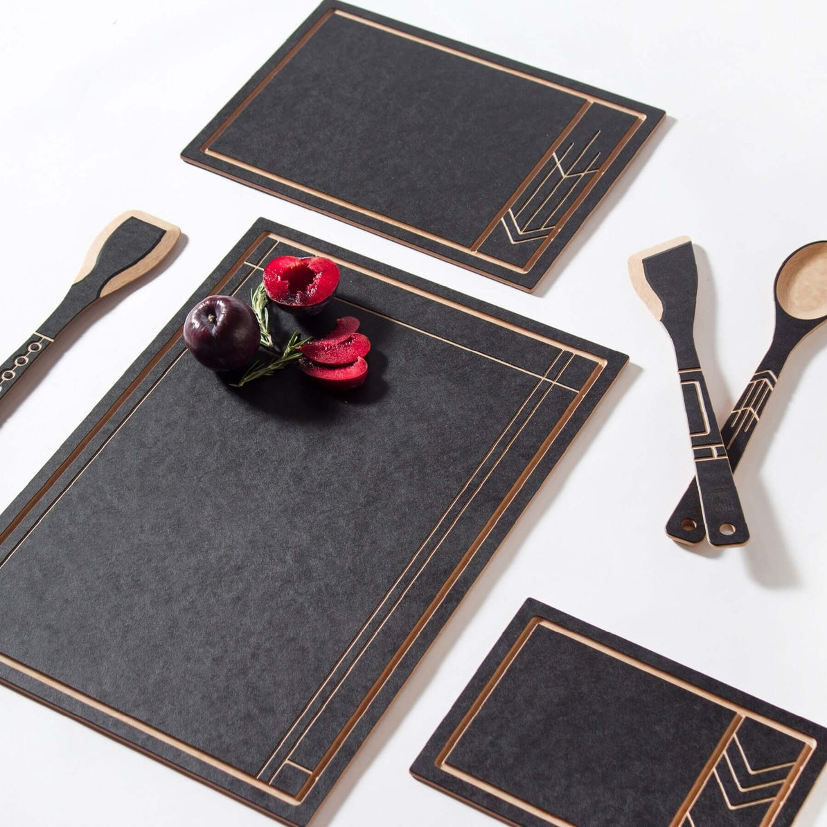 A black cutting and serving board with a geometric pattern in a Wright-style design, accompanied by matching black serving utensils, presented with a dish of fruit on top.