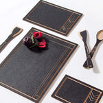 A black cutting and serving board with a geometric pattern in a Wright-style design, accompanied by matching black serving utensils, presented with a dish of fruit on top.