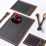 A black cutting and serving board with a geometric pattern in a Wright-style design, accompanied by matching black serving utensils, presented with a dish of fruit on top.
