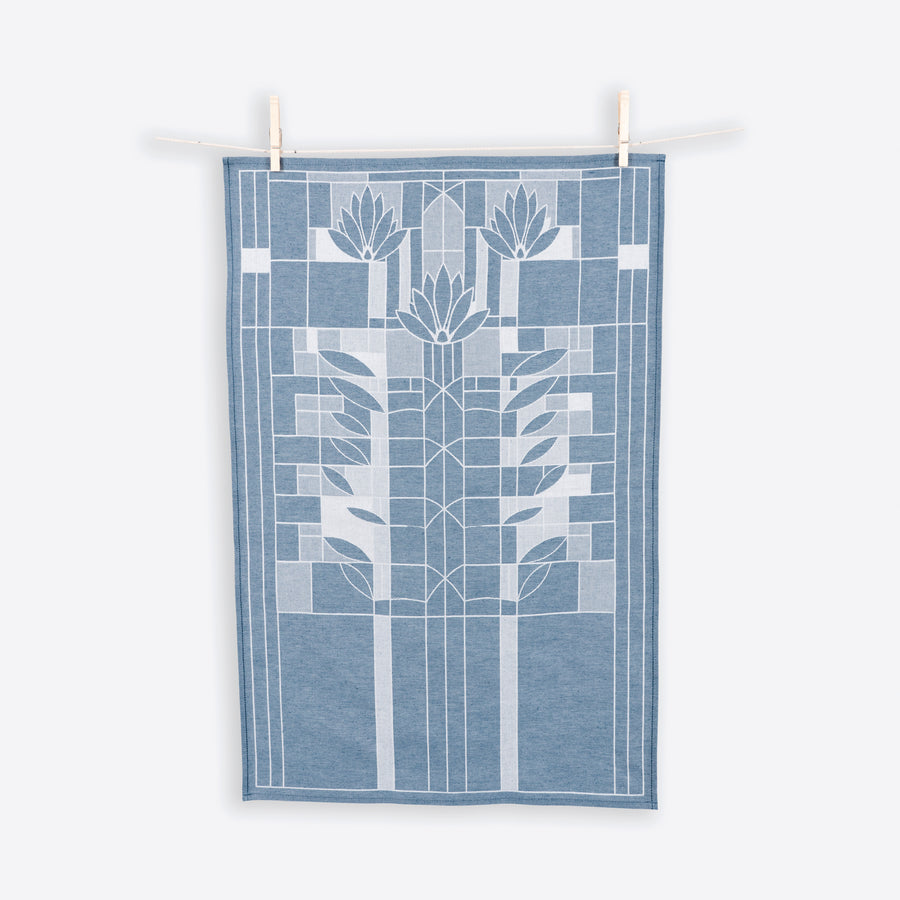 A blue and white cotton tea towel with a geometric waterlilies pattern hanging on two wooden clothespins against a white background.