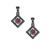 A pair of earrings featuring a geometric design with a red bead in the center, plated in hematite, on a white background.
