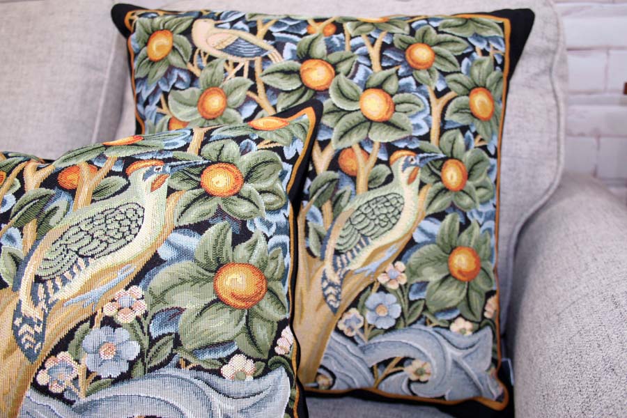 Two square-shaped throw pillows with a floral and bird design, in green, blue, and brown colors, on a sofa.